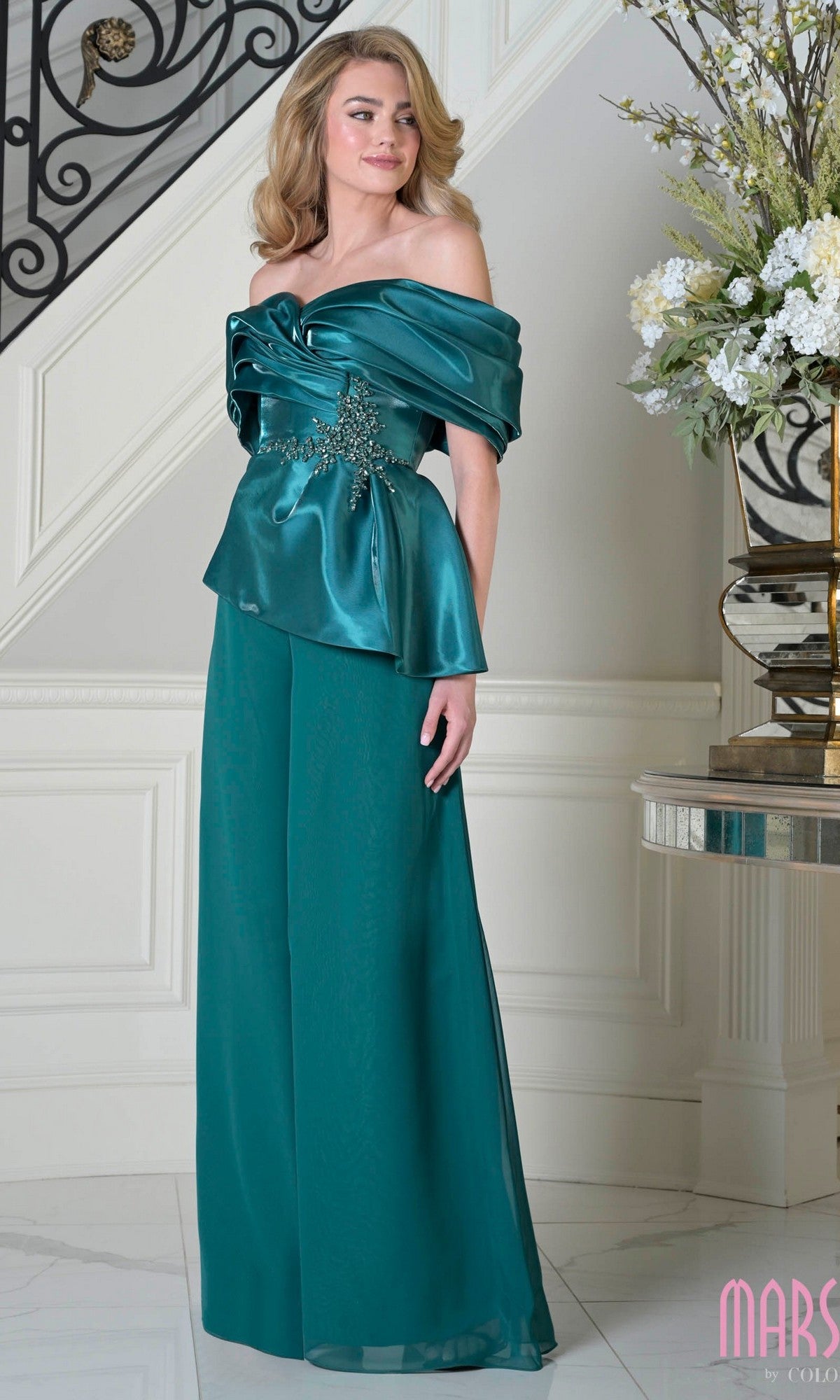 Woman stands indoors in the elegant Long Formal Dress: Marsoni MV1410 in deep green, featuring an off-the-shoulder bodice and wide leg chiffon pants, beside a silver table with a floral arrangement.